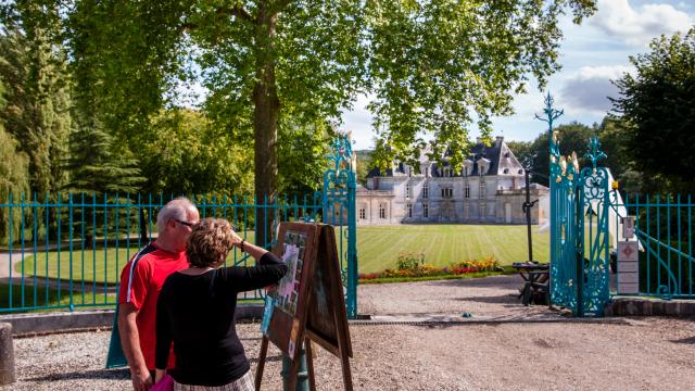 Photo d'un couple devant l'entrée du parc du château d'Acquigny avec sa barrière en fer forgée bleu, ses arbres feuillus et ses fleurs